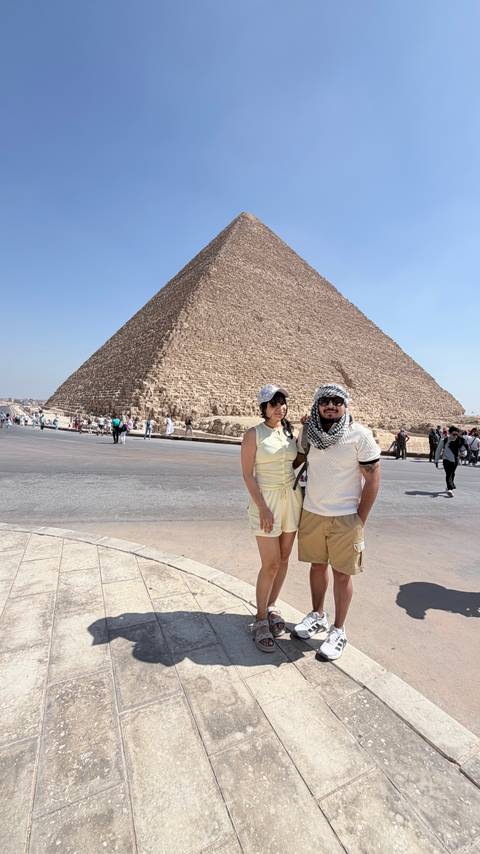       Smiling couple poses in front of the Great Pyramid of Giza on a bright, cloudless day while other tourists explore the site.
  