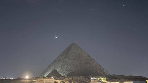       Nighttime silhouette of a pyramid at Giza beneath a crescent moon and star-dotted sky with faint ground lighting.
  