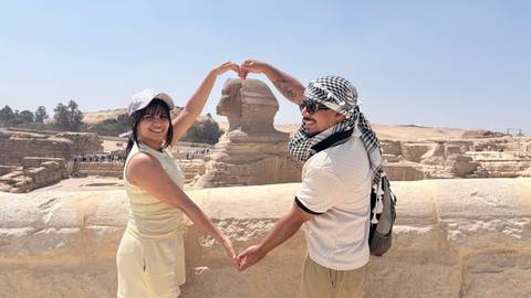       Couple makes a heart shape with their arms while posing in front of the Sphinx on the sunny Giza Plateau.
  