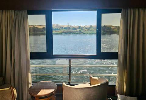       Panoramic Nile view through cruise cabin window with armchairs in foreground.
  