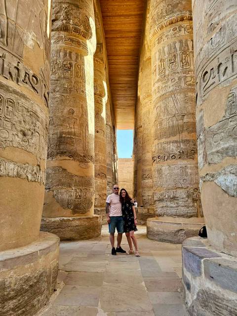       A smiling couple stands among the massive carved columns of Karnak Temple with blue sky above.
  