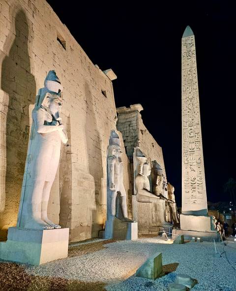       Illuminated statues and an obelisk tower over Luxor Temple courtyard at night.
  