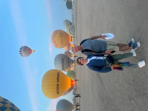       Couple posing on desert launch field with colorful hot-air balloons ascending at dawn.
  