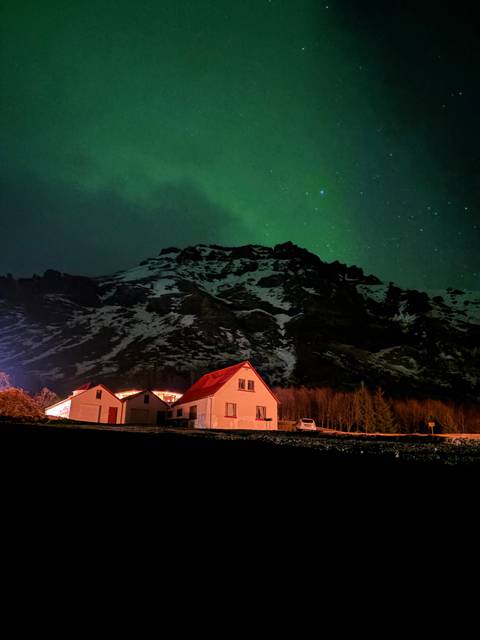       Northern lights glow green above a snow-tipped mountain and a cozy red-roofed farmhouse at night.
  