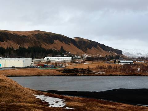       An Icelandic river runs past low industrial buildings and brown hills on a grey day.
  