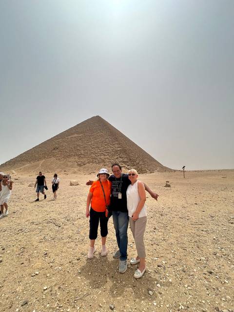       Three tourists pose before a desert pyramid while others explore the sandy plateau.
  