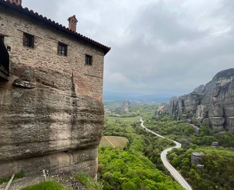       Sweeping valley view of lush Meteora landscape with rock pillars and winding road.
  