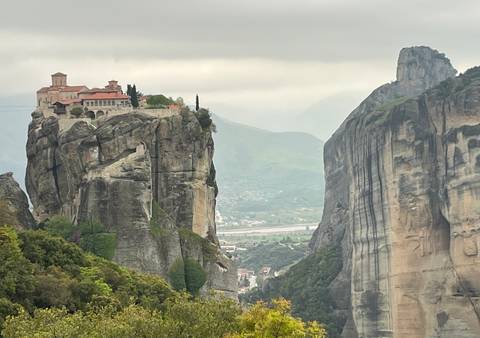       Iconic Meteora monastery balanced on narrow rock column against misty mountainous backdrop.
  