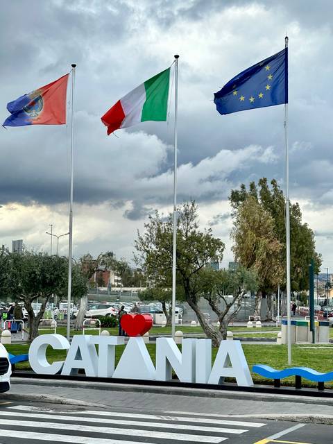       Flagpoles stand in a parking area with cloudy skies and parked cars behind.
  