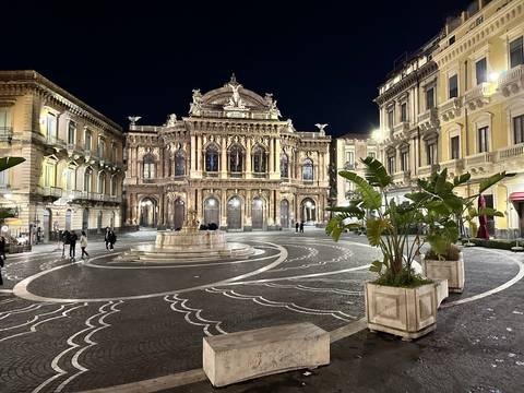       Illuminated baroque theatre façade overlooking a patterned piazza at night in Catania.
  