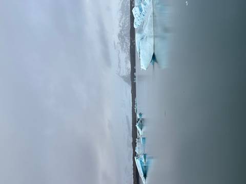       Calm glacial lagoon with floating icebergs and snow-covered mountains beneath a grey overcast sky
  