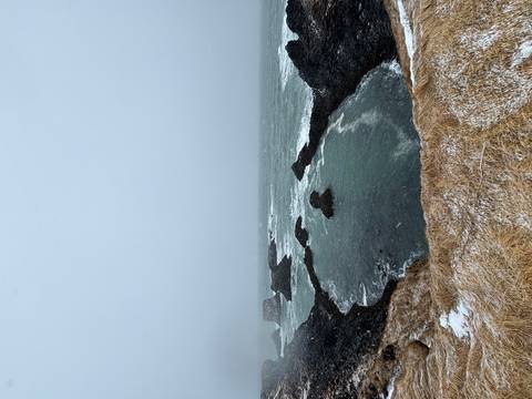       Snow flurries sweep across rugged sea cliffs and churning turquoise water beneath a grey sky
  