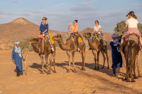       Group of tourists ride camels led by guides across a sandy desert plain with mountains in the background.
  