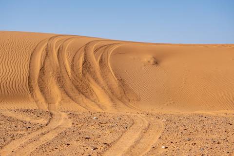       Curving tire tracks carve into smooth desert dunes under a clear pale blue sky.
  