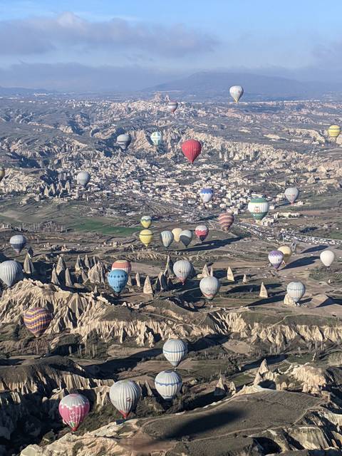       High aerial shot of dozens of hot-air balloons drifting over rocky spires and the town in Cappadocia.
  
