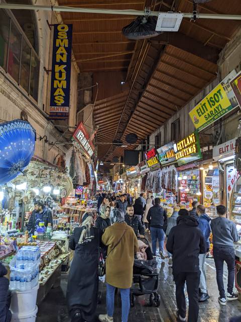       Bustling indoor bazaar with hanging goods, bright neon signs and crowds of shoppers in a narrow market aisle.
  