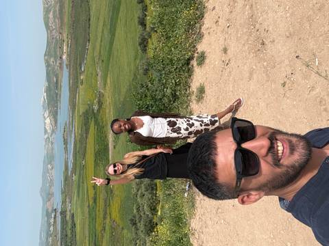       Three travellers smiling for a selfie at a hilltop viewpoint with a lake, rolling green hills and distant mountains behind them.
  