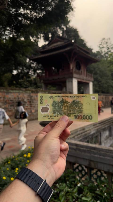       Hand holding a 100,000 VND banknote aligned with historic temple courtyard in background
  