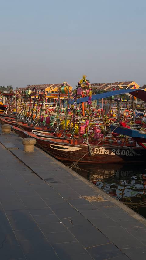       Row of wooden tour boats festooned with colorful lanterns moored along a riverside at sunset
  