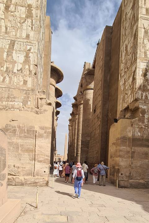       Row of monumental sandstone columns covered in hieroglyphs leading to distant obelisk under bright sky
  
