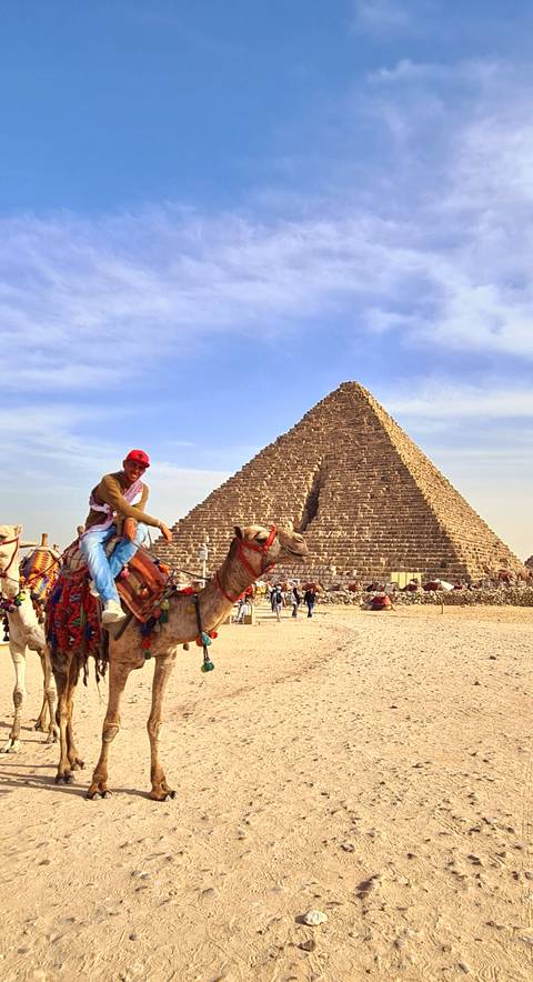       Traveler riding a camel in front of a large pyramid on a sunny desert day
  