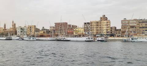       Riverside town with moored sailboats and weathered apartment blocks seen across choppy water
  