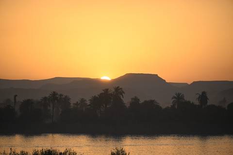       Golden sun setting behind layered desert hills silhouetting palm trees along a calm river.
  