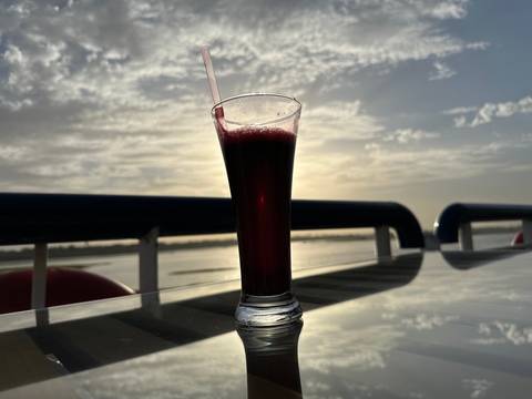       Tall glass of dark juice on a reflective deck table with soft sunset light over a river in the background.
  