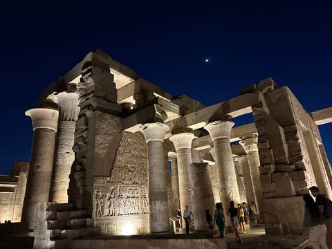       Beautifully lit columns and carvings of Kom Ombo Temple under a dark blue night sky with a crescent moon.
  