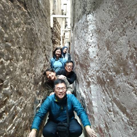       Five travellers squeeze through a very narrow, rough-stone alley, smiling and posing for the camera.
  