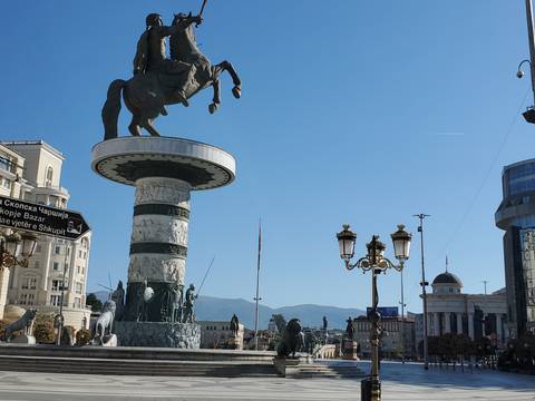       Large equestrian statue atop an ornate pedestal dominates a modern city square under a clear blue sky.
  