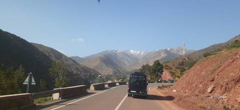       A minibus drives along a winding mountain highway with snow-capped Atlas peaks in the distance and red earth slopes nearby.
  