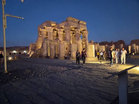       Nighttime view of a flood-lit Egyptian temple with tourists walking on the stone forecourt.
  