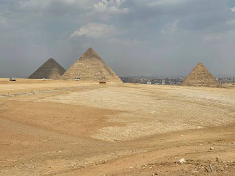       Wide desert panorama of the three Pyramids of Giza rising over the sandy plateau with distant city haze.
  