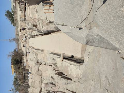       Granite trench of the Unfinished Obelisk in Aswan quarry with scattered visitors walking along the barrier.
  