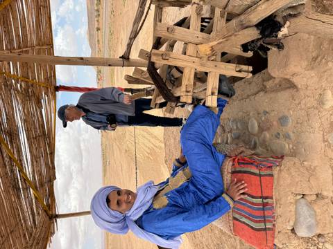       A Berber man relaxes under a rustic desert shelter while a visitor looks on.
  