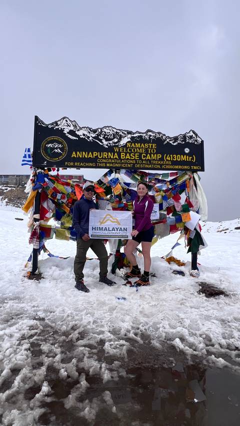       Two trekkers pose by the colourful prayer-flag-covered sign at Annapurna Base Camp in snowy surroundings.
  