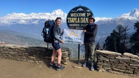       Trekkers smile beside a 'Welcome to Thapla Danda' sign with panoramic snowy peaks behind.
  