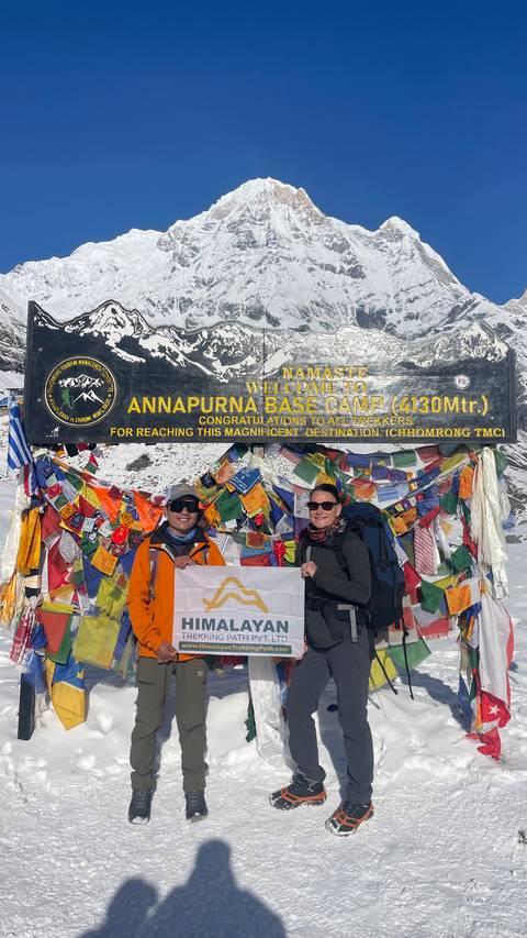       Close-up of two trekkers holding a banner in front of the Annapurna Base Camp welcome board covered in prayer flags.
  