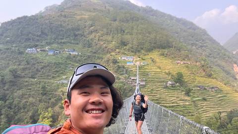       Smiling selfie on a long suspension bridge over a verdant river valley with rice terraces.
  