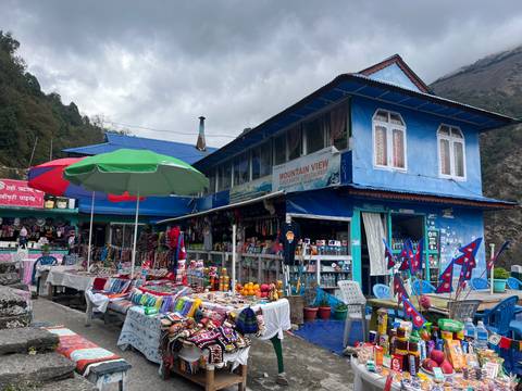       Colourful mountain village bazaar with blue guesthouse, souvenir stalls and umbrellas under an overcast sky.
  