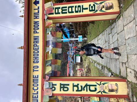       Trekker stands beneath a welcome arch to Ghodepani Poon Hill holding a camera with lodges in the background.
  