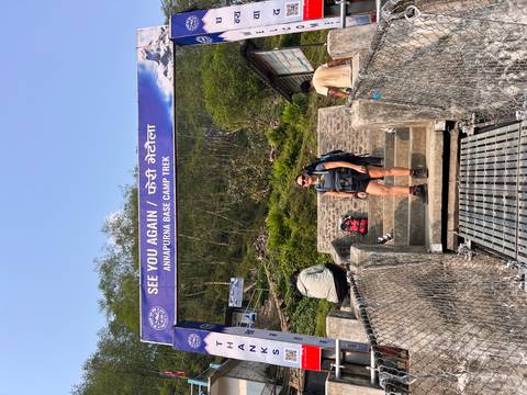       Trekker pauses under a farewell banner reading 'See You Again / Annapurna Base Camp Trek' at a suspension bridge exit.
  