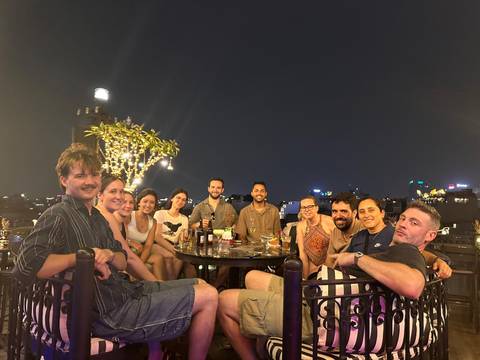       Smiling travellers share drinks around a rooftop table at night with a city skyline twinkling behind them.
  