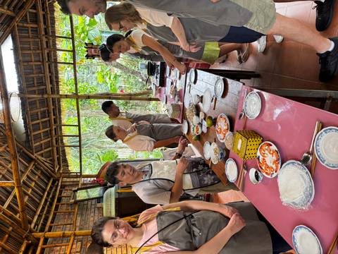       Travellers in aprons line a long table preparing ingredients during a hands-on Vietnamese cooking class in an open-air kitchen.
  