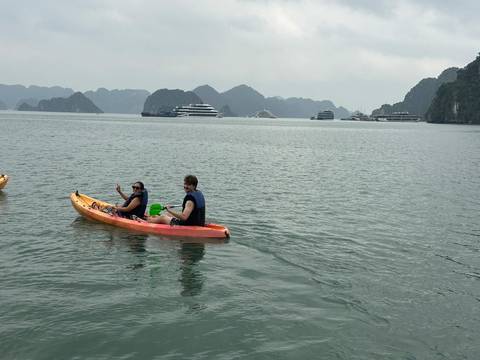       Two travelers paddle an orange kayak on the calm emerald waters of Halong Bay with cruise ships afar.
  