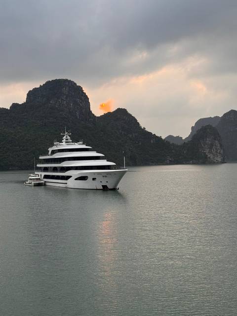       White luxury cruise ship anchored in Halong Bay at sunset with limestone peaks silhouetted.
  