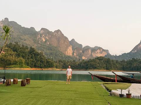       A traveler stands on lush grass beside emerald Cheow Lan Lake framed by sheer limestone cliffs in Khao Sok.
  