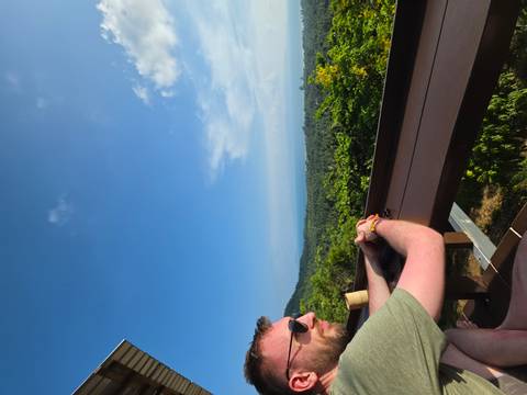       Man wearing sunglasses gazes over a vast forested coastline from a hilltop café viewpoint.
  