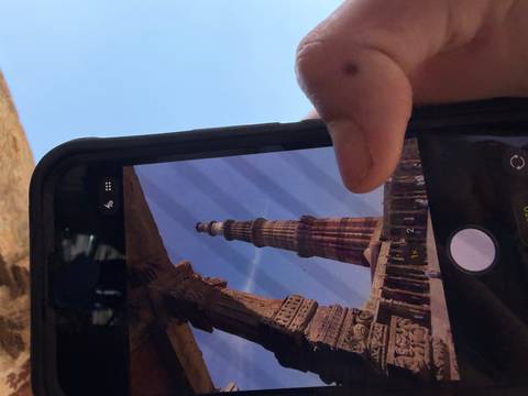       Close-up of a smartphone photographing the soaring Qutub Minar through stone archways.
  